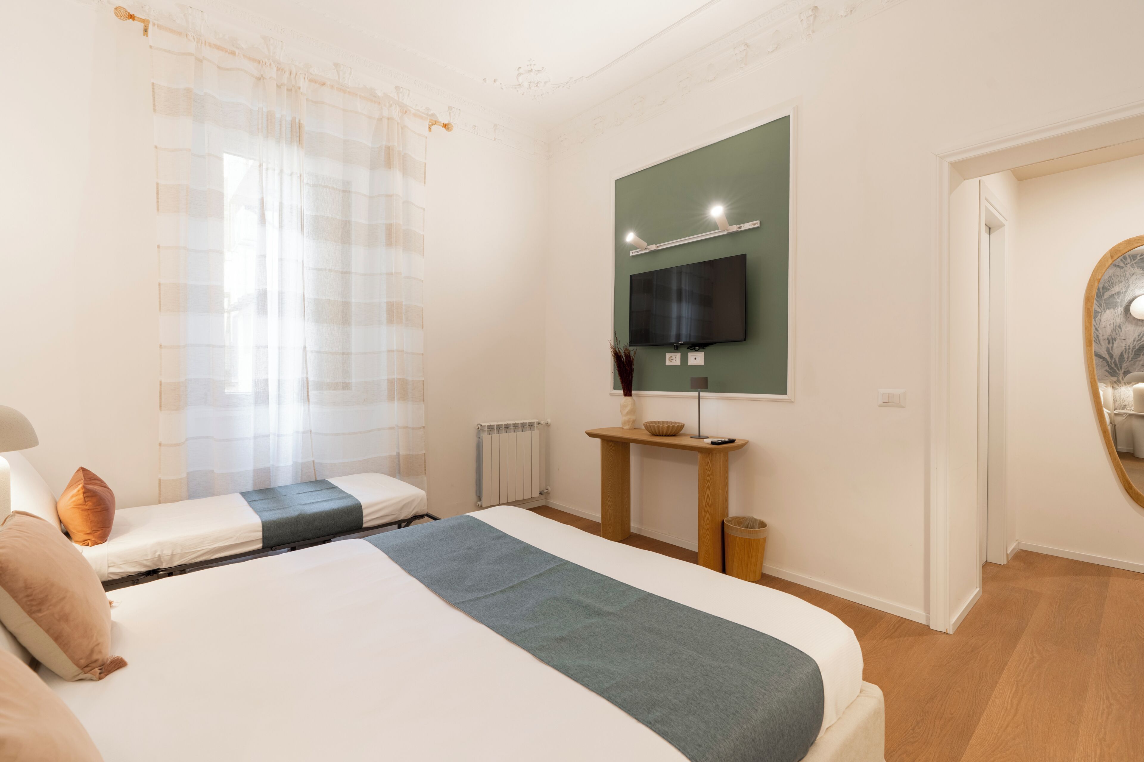 A bedroom with a bed, a bedside table, a mirror, a television, and a window with curtains.