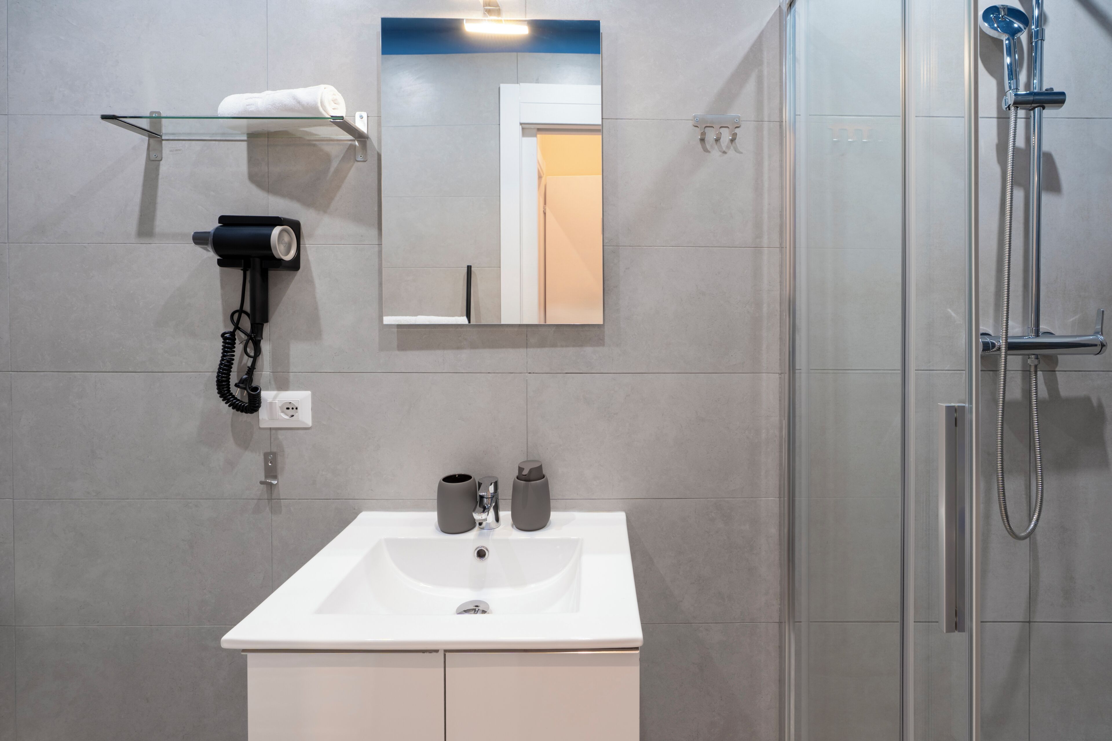 Un baño con lavamanos blanco, espejo, toallero, secador de pelo y ducha.