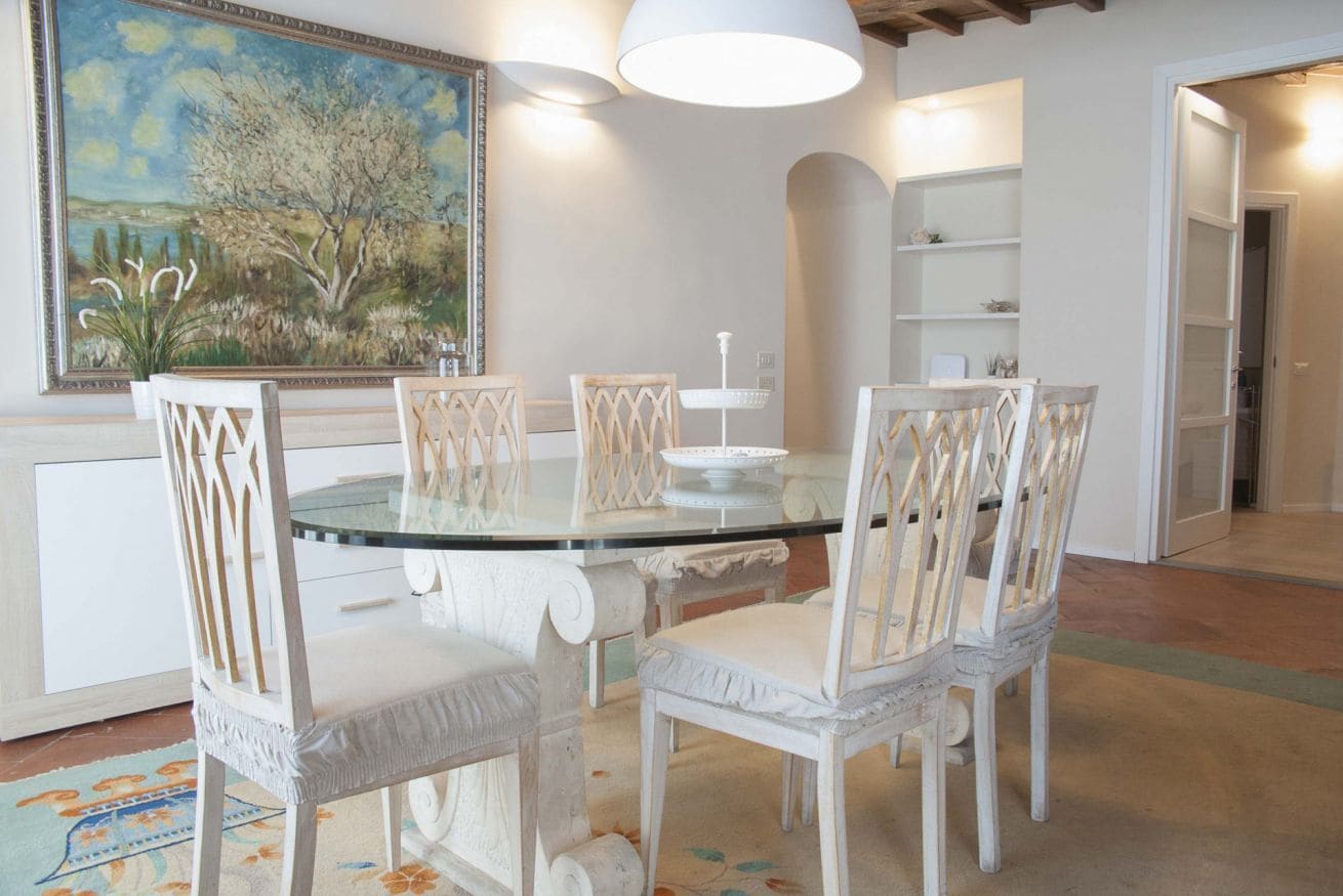 A dining area with a glass table surrounded by white chairs with intricate back designs, a painting on the wall, and a white pendant light above.