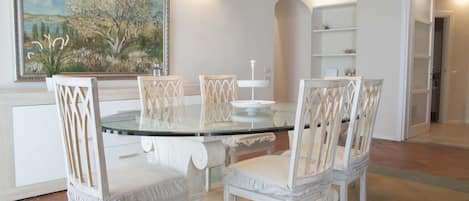 En matplats med ett glasbord omgivet av vita stolar med detaljerade ryggstöd, en tavla på väggen och en vit hänglampa ovanför.