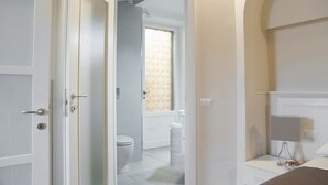 Habitación de hotel con baño visible a través de una puerta abierta, una cama, una mesita de noche y un revistero.