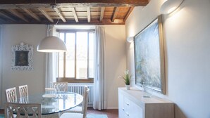 Un comedor con una mesa de vidrio, sillas, una lámpara colgante, un radiador y una ventana con cortinas.