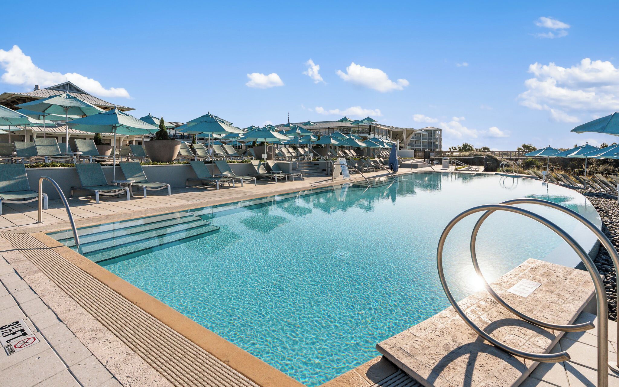 A clear swimming pool with lounge chairs and umbrellas, surrounded by buildings.