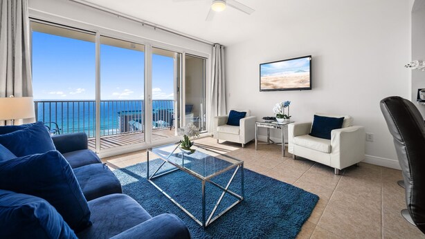 A modern living room with a large glass table, two white armchairs, a blue sofa, and a view of the ocean through large sliding doors. - Ariel Dunes I 1906 *3 pools *Beach access* (Miramar Beach)