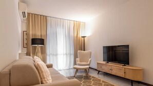 Ein modernes Hotelzimmer mit einer Couch, einem Sessel, einem Fernseher auf einer Holzkommode und einem großen Fenster mit Vorhängen.