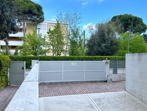 A paved outdoor area with a metal fence, a utility box, and a hedge. - COND. PELICAN - B2 4+1 - B50C2T (Bibione Pineda)
