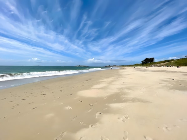 Une vaste plage de sable fin parsemée d’empreintes, un ciel dégagé et des bâtiments qui se dessinent à l’horizon.