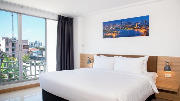 A modern hotel room with a large bed, a cityscape painting, and a view of buildings and greenery through the window.