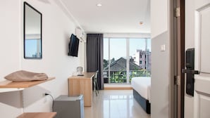 A hotel room with a bed, a desk, a chair, a small fridge, a mirror, and a view of buildings outside.