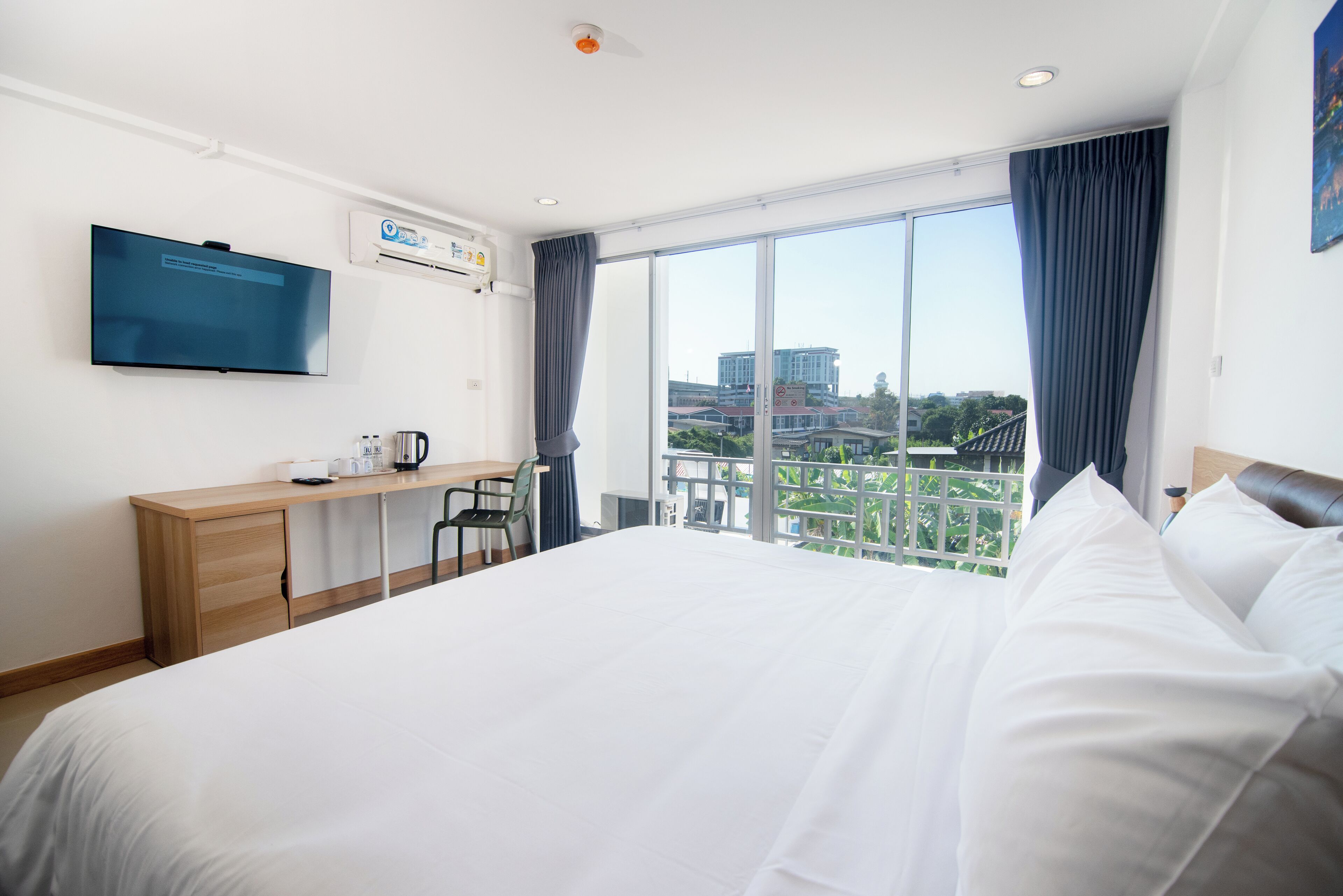 A hotel room with a large bed, a desk with a chair, a flat-screen TV, and a window with a view of buildings.