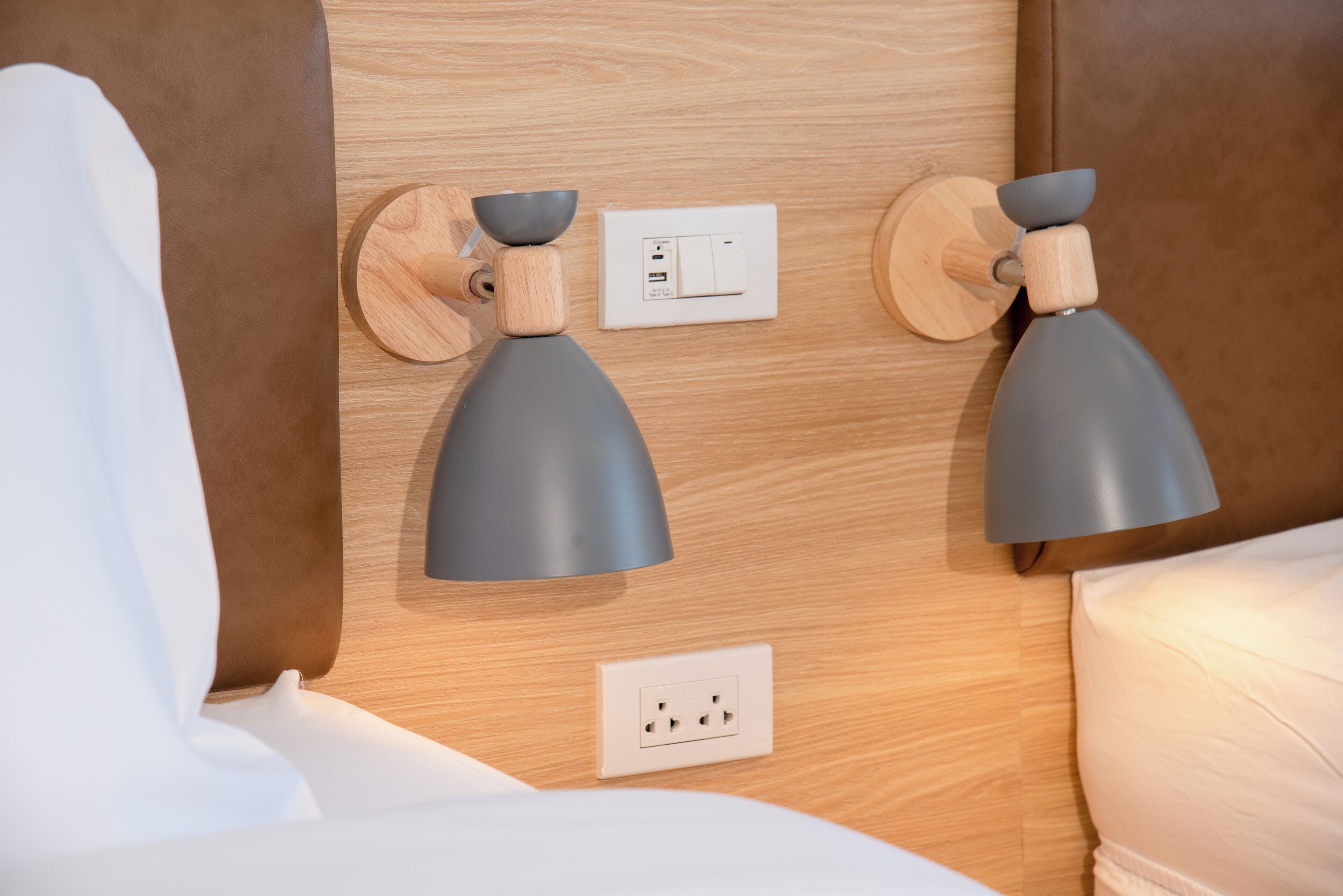 A modern room with a wooden headboard, two wall-mounted lamps, a power outlet, and a bed with white bedding.