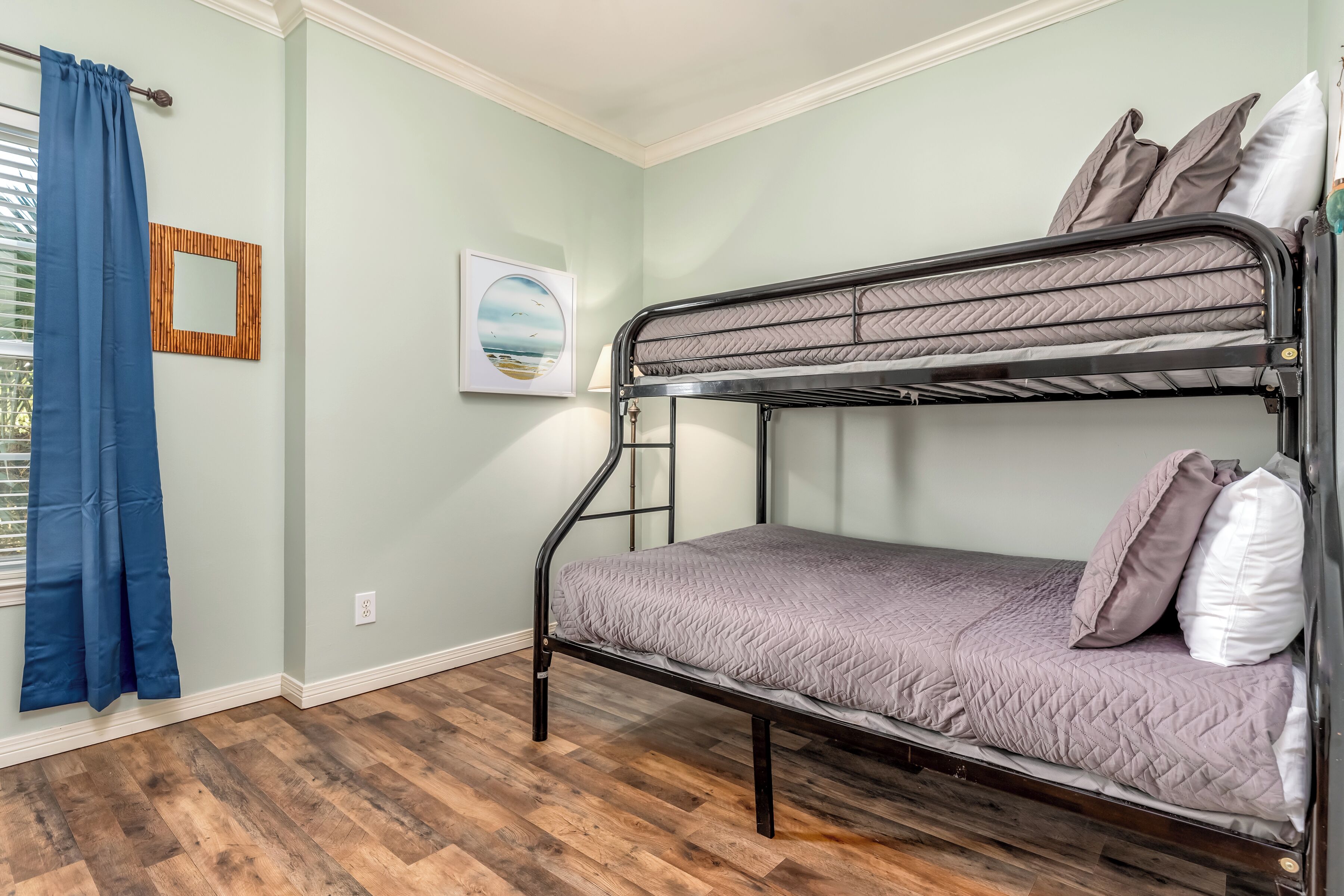 A bunk bed in a room with light green walls, a wooden floor, a framed picture, and a blue curtain.