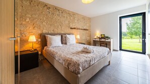 Ein Schlafzimmer mit Steinwand, einem Bett mit floralem Überwurf, zwei Nachttischlampen, einem Schreibtisch und Blick auf eine grüne Landschaft durch eine Glastür.