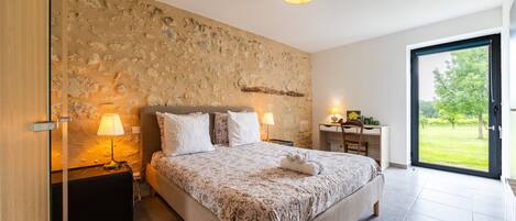 A bedroom with a stone wall, a bed with a floral bedspread, two bedside lamps, a desk, and a view of a green landscape through a glass door.