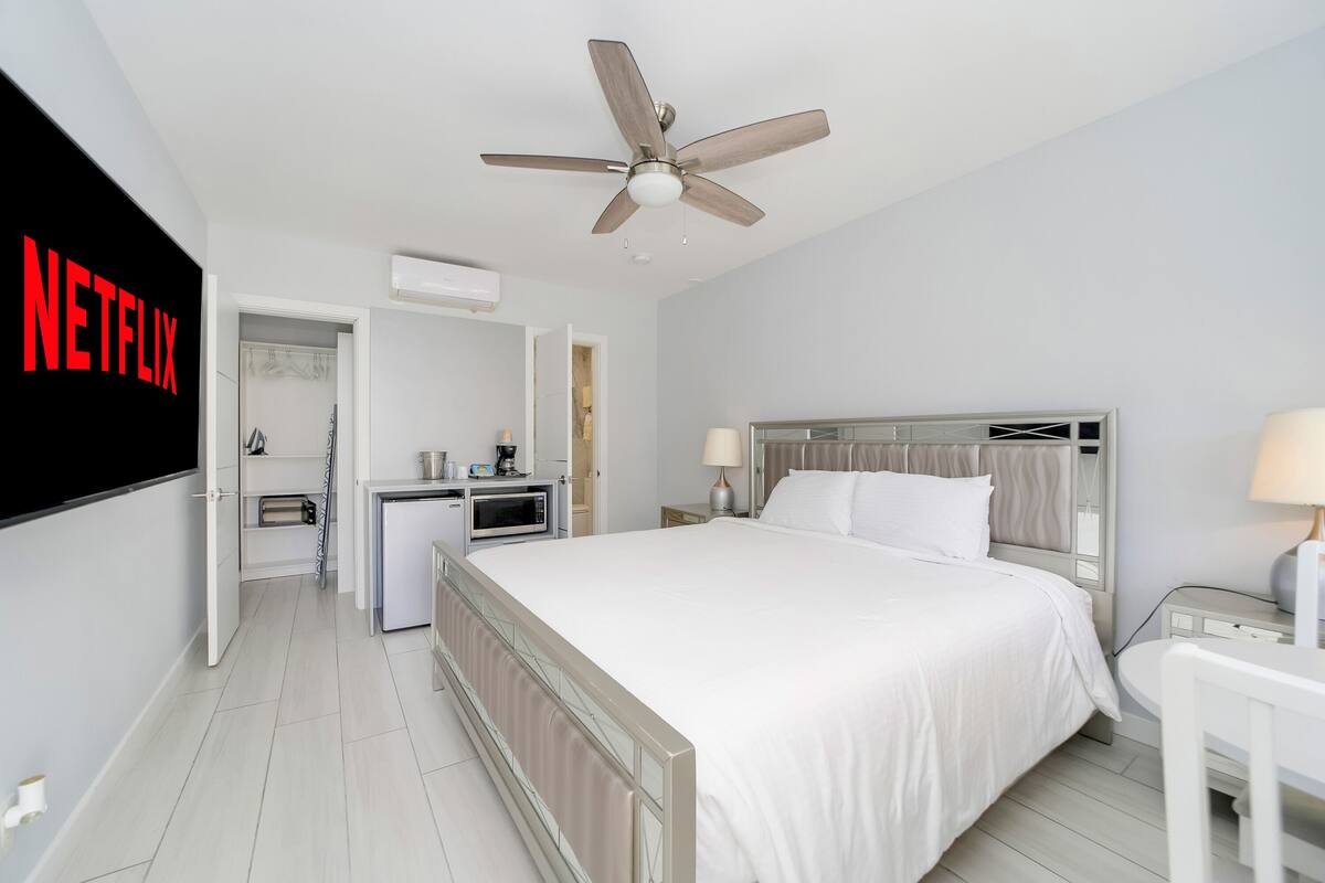 A hotel room with a bed, bedside table, lamp, ceiling fan, and a kitchenette area with a microwave and coffee maker.