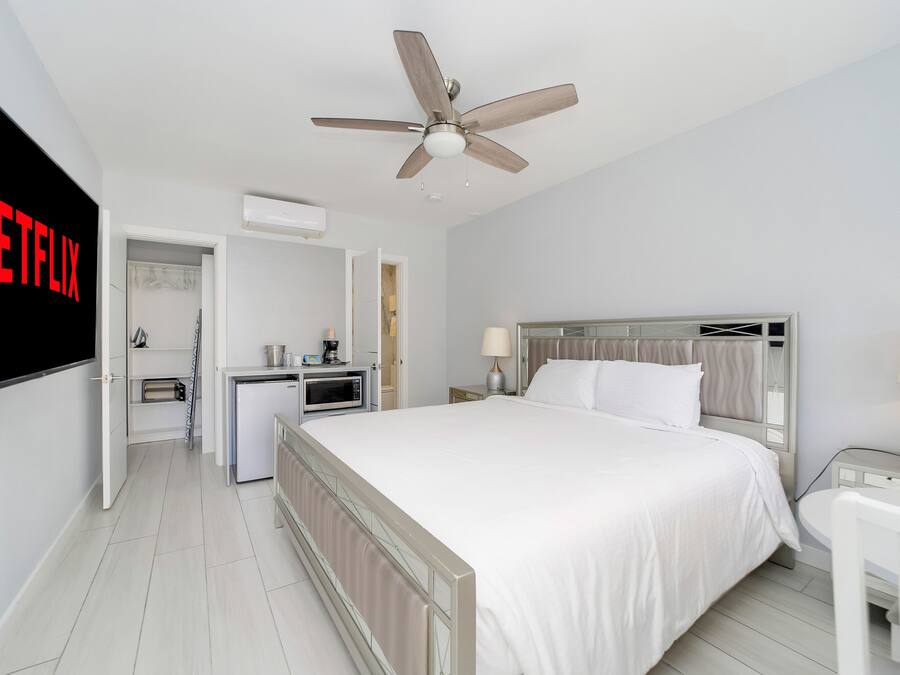A hotel room with a bed, bedside table, lamp, ceiling fan, and a kitchenette area with a microwave and coffee maker.