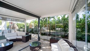 A modern outdoor patio with wicker furniture, glass tables, and a view of a lush garden and building in the background. - Deco Boutique Hotel (Fort Lauderdale)