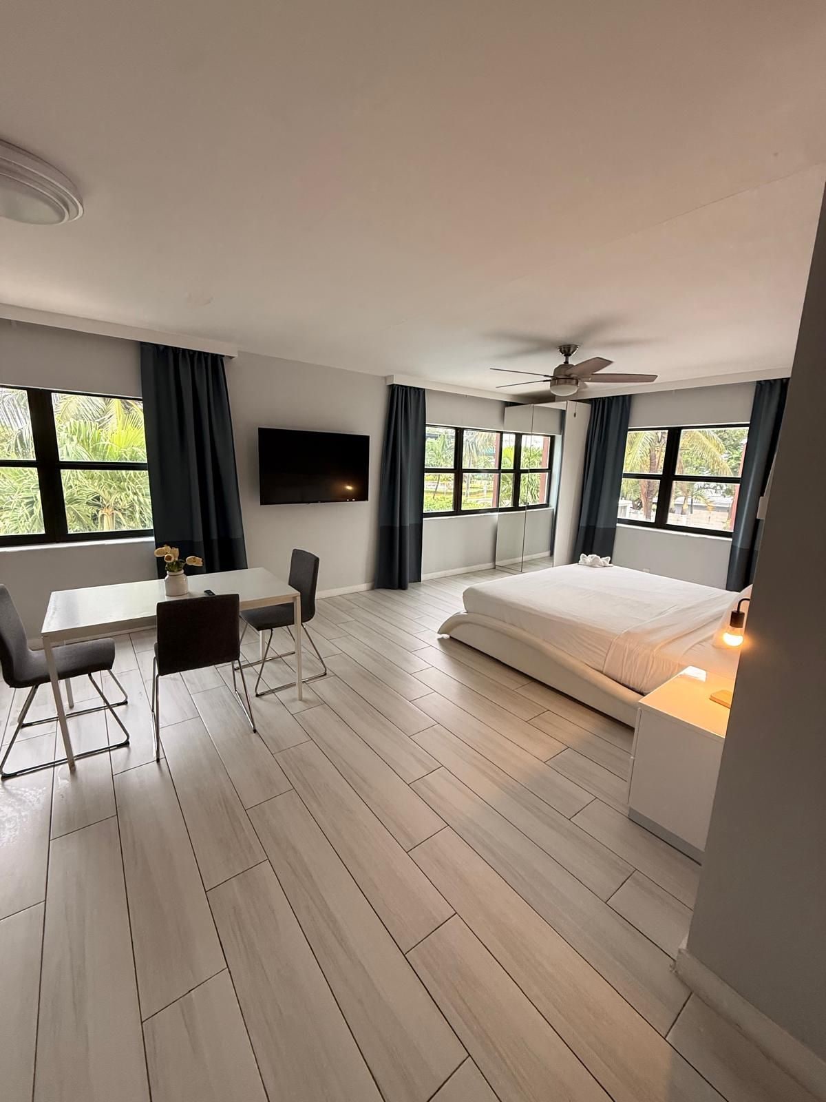 Een moderne hotelkamer met een bed, bureau, stoelen, een flatscreen-tv en grote ramen met gordijnen.