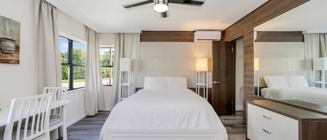 A modern bedroom with a large bed, white furniture, a ceiling fan, and a view of greenery outside.