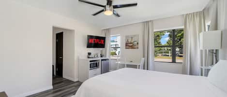 Habitación de hotel con una cama grande, ventilador de techo, televisor mostrando 'NETFLIX', kitchenette y vistas al exterior a través de una ventana.