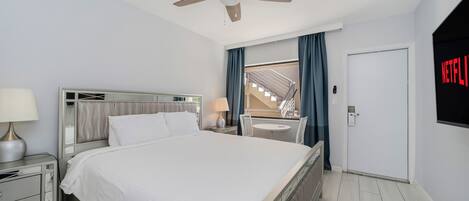A modern hotel room with a large bed, a ceiling fan, a TV mounted on the wall, and a view of the outside balcony.