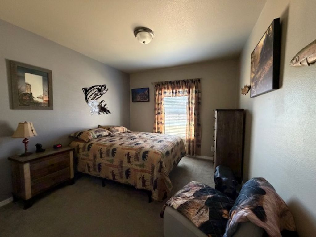 A bedroom with a bed, a dresser, a nightstand, a lamp, a window with curtains, and a wall-mounted artwork.