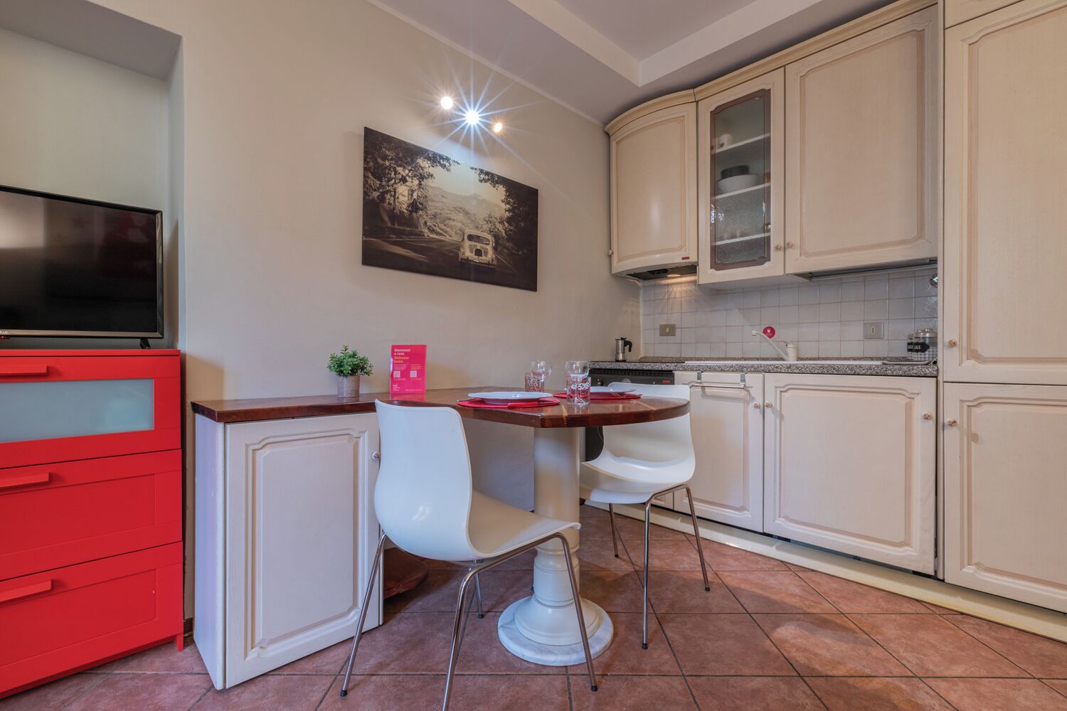Une cuisine de taille réduite avec des meubles blancs, un meuble rouge, une table à manger avec deux chaises blanches, une petite plante en pot et un tableau accroché au mur.