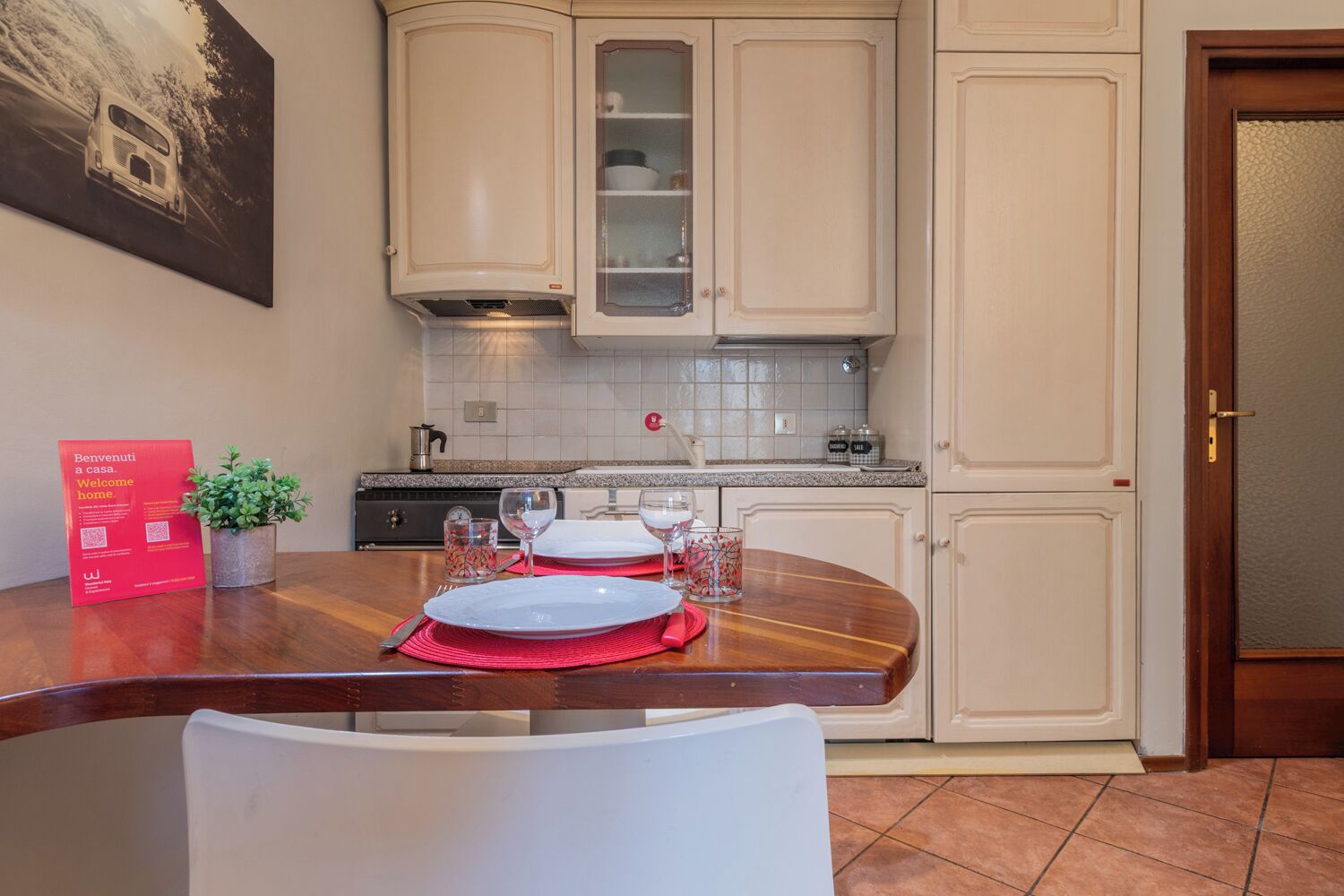 Un coin repas avec une table en bois dressée pour deux personnes, une plante en pot et un panneau rouge indiquant « Bienvenue ».