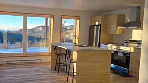 A modern kitchen with wooden cabinets, a stainless steel refrigerator, and a large window offering a view of a snowy landscape.