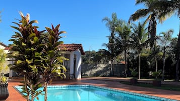 Piscina com borda azulejada, cercada por palmeiras e um pequeno edifício com telhado vermelho.