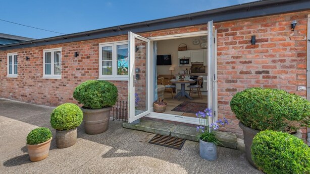 A brick building with a door open to a dining area, potted plants, and a clear sky. - 1 Bed in Ashton (oc-p30484) (Ashton)