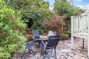 A stone patio with a table and chairs, surrounded by dense foliage and a white fence. - 2 Bed in Polruan (oc-g30534) (Fowey)