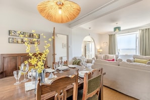 Ein Essbereich mit einem Holztisch, Stühlen und einer Vase mit gelben Blumen. Im Wohnbereich befindet sich eine Couch und ein Fenster mit Vorhängen.