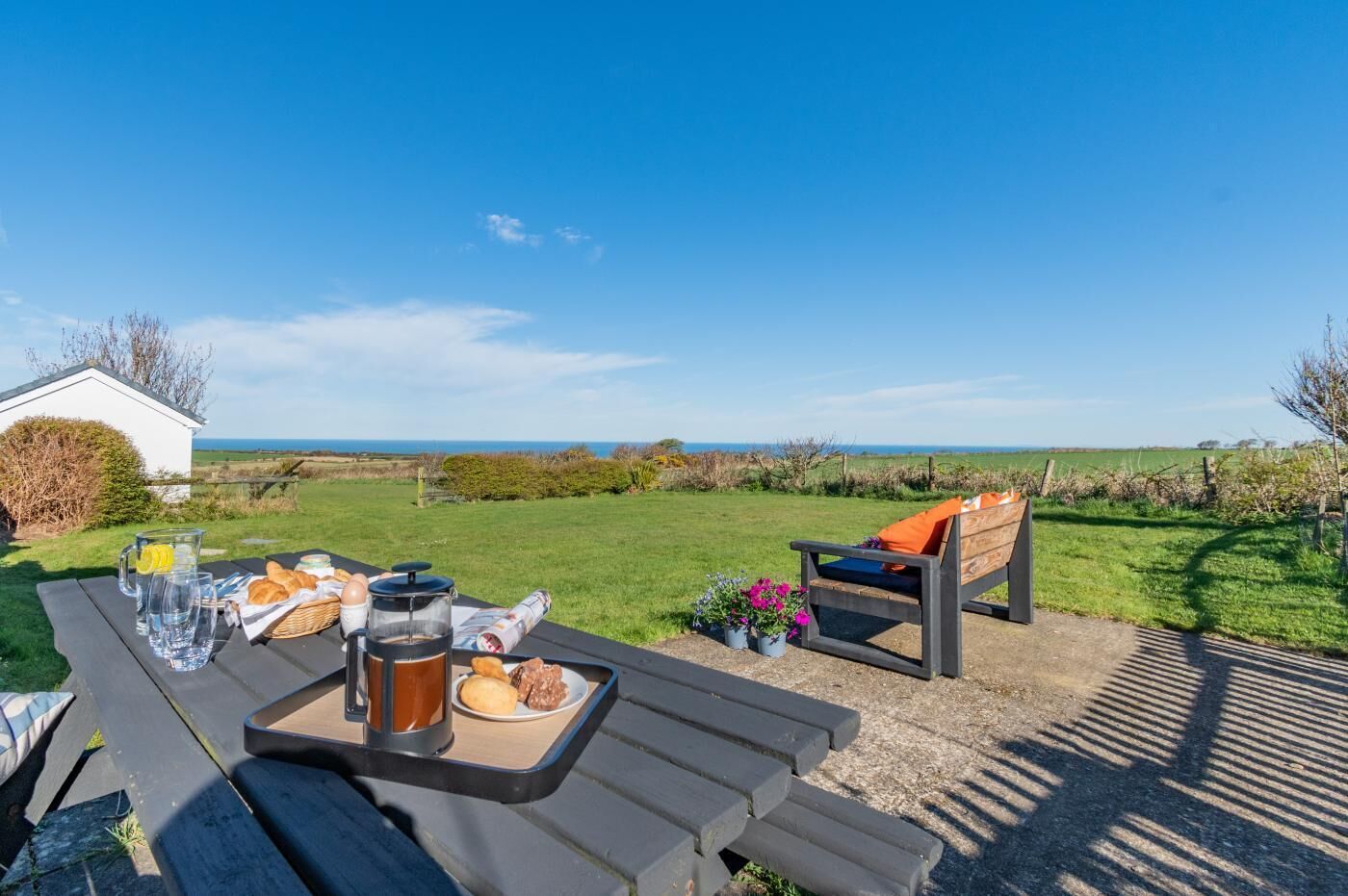 An outdoor dining area with a view of the sea, a picnic table with food and drinks, and a bench with cushions.