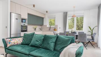 Een moderne keuken met een koelkast, oven en magnetron. Er is een eethoek met een tafel en stoelen. Een groene bank met een bloemenkussen bevindt zich op de voorgrond.