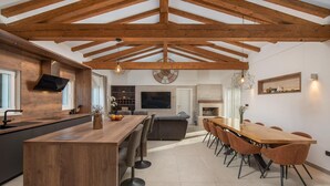 Une cuisine moderne avec un îlot central en bois, un coin repas avec une longue table et des chaises, et un salon avec un canapé et une télévision.