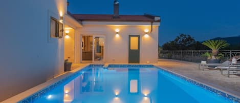 A swimming pool with a clear blue edge, a small building with a door and windows, and a patio area with outdoor furniture.