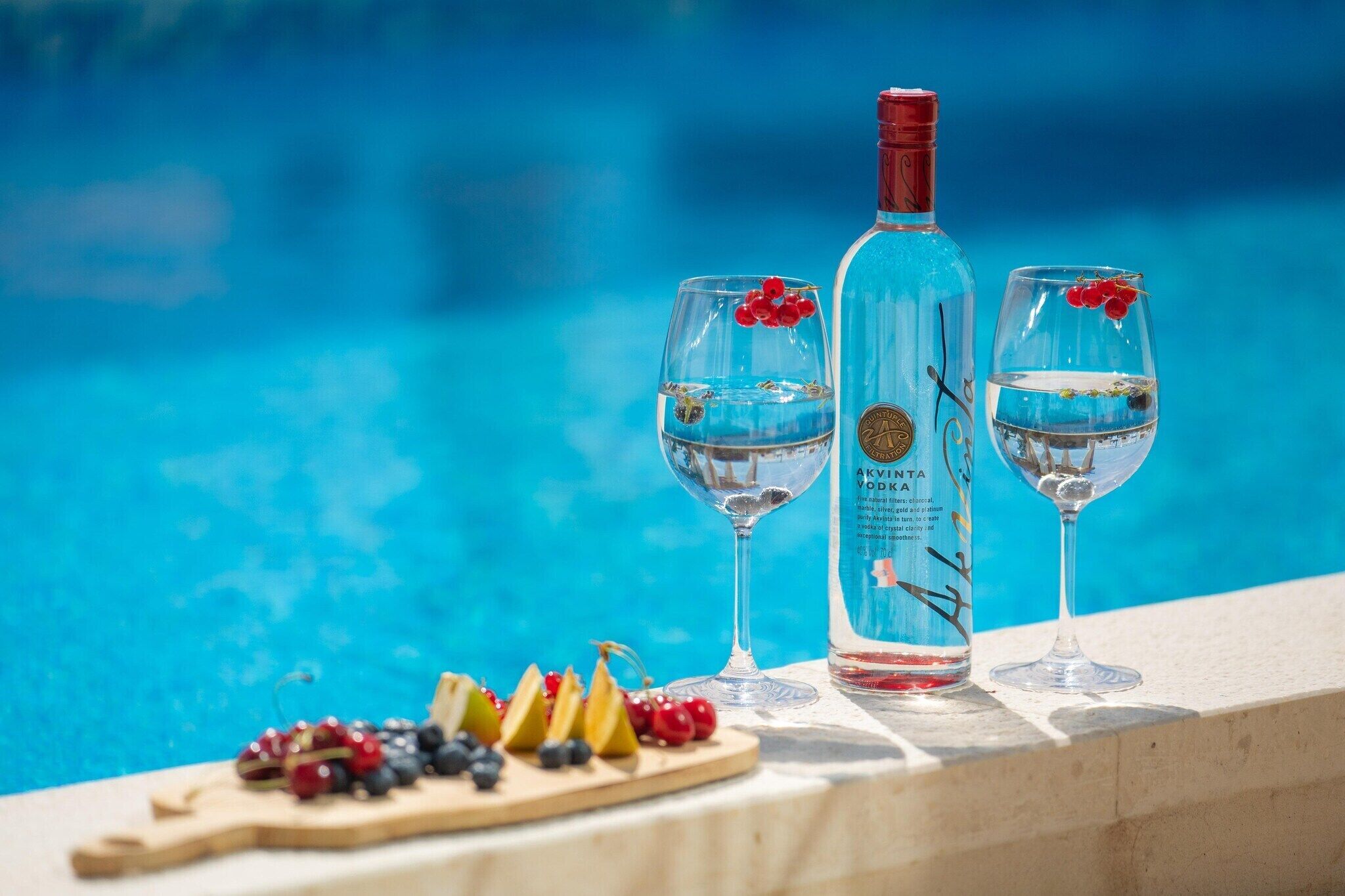 Una botella de vodka Akvinta y dos vasos sobre una mesa junto a la piscina, con frutas.