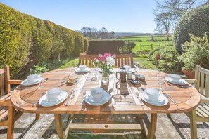 A wooden table set for six with white plates, bowls, and cutlery, surrounded by chairs, set against a backdrop of a lush garden and rolling hills. - 4 Bed in North Tamerton (oc-h30361) (North Tamerton)