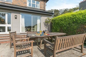 Eine Terrasse mit Holztisch und -stühlen, einer Glastür und einer Hecke.