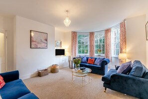 Ein Wohnzimmer mit zwei Sofas, einem Fernseher, einer Deckenleuchte und großen Fenstern mit Vorhängen.