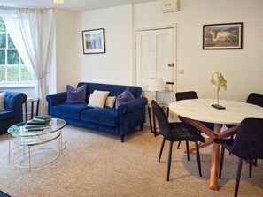 Ein Wohnzimmer mit blauem Sofa, einem Glastisch, einem runden Esstisch mit Stühlen und zwei gerahmten Bildern an der Wand.