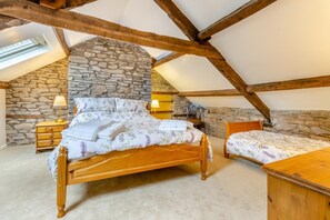 A bedroom with a stone wall, wooden beams, a large bed, a smaller bed, a nightstand, a lamp, and a wooden dresser. - 6 Bed in Lower Chapel (oc-c30673) (Lower Chapel)