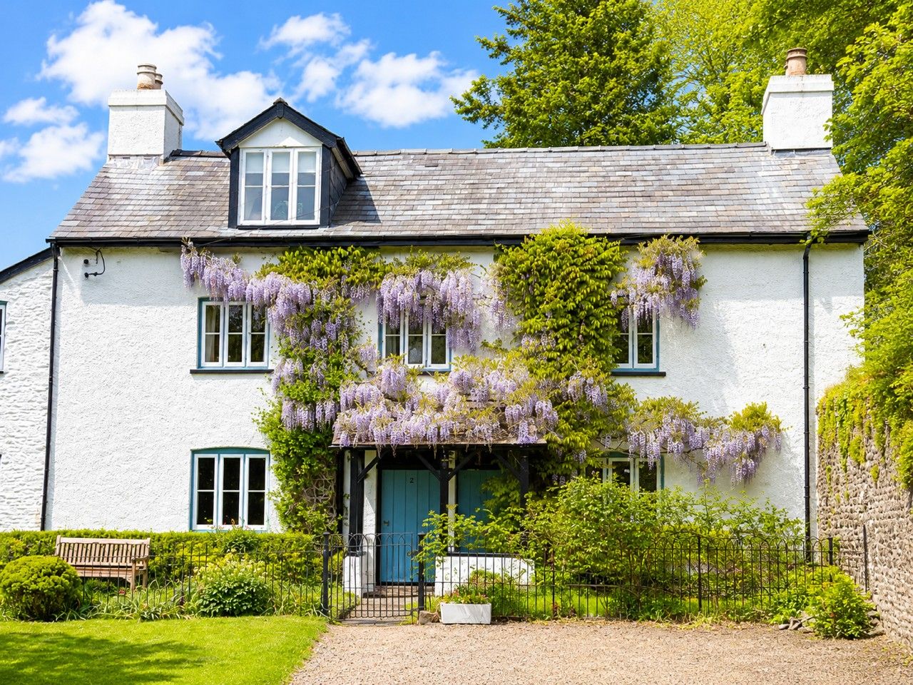 A white house with a blue door, surrounded by greenery and a gravel path.