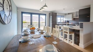 A modern kitchen with a dining area, featuring a large window, a clock, and a dining table set for four. - 5 Bed in Bwlchtocyn (oc-s30770) (Bwlchtocyn)