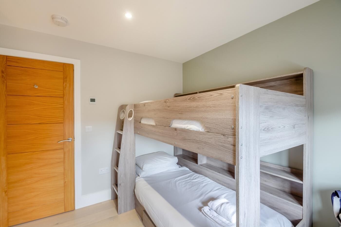 Uma cama beliche com cabeceira de madeira e uma cama de solteiro embaixo. Há uma porta de madeira e um interruptor de luz na parede.