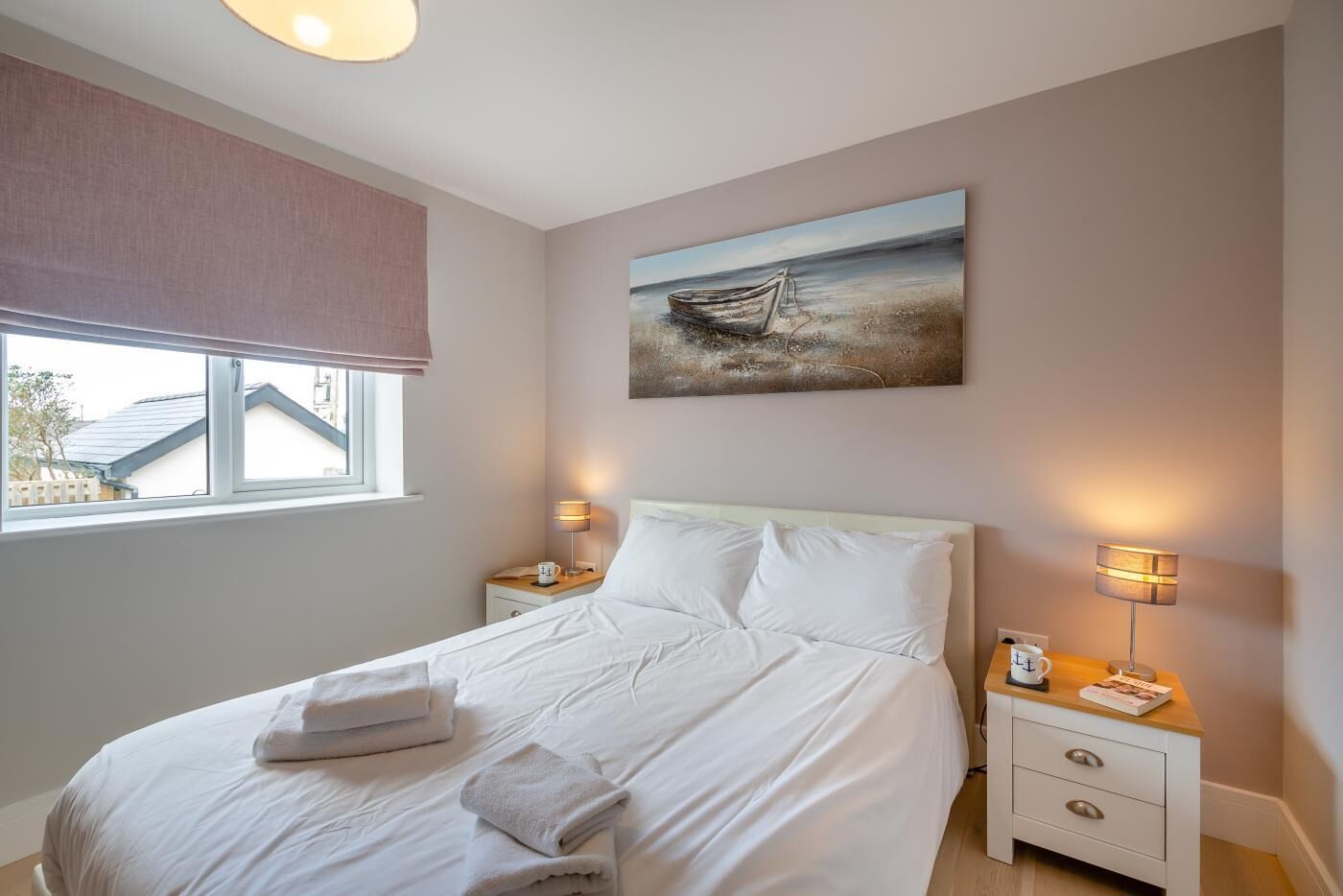 A neatly made bed with white linens and folded towels, a bedside table with a lamp, and a painting of a boat on the wall.