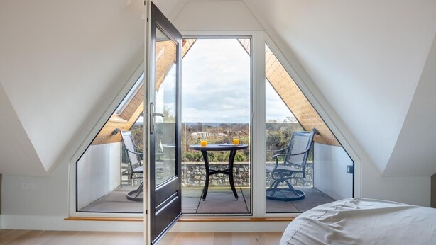 A room with a sloped ceiling, a sliding glass door leading to an outdoor patio with a table and chairs, and a bed with a white cover. - 5 Bed in Bwlchtocyn (oc-s30770) (Bwlchtocyn)