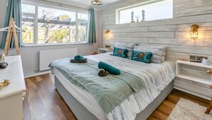 Een slaapkamer met een groot bed, houten vloeren, een raam met gordijnen en een nachtkastje met een lamp.