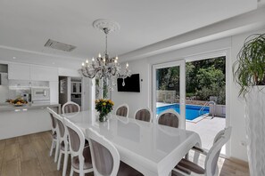 Una cocina moderna con armarios blancos, un comedor con mesa y sillas blancas, una araña y vista a una piscina.